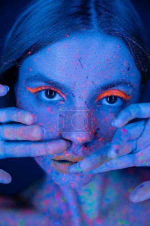 Téléchargez les photos : Portrait de femme tenant les mains près du visage avec maquillage au néon et peinture fluorescente isolée sur bleu foncé - en image libre de droit