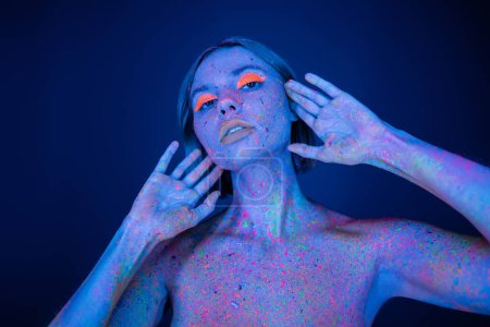 Téléchargez les photos : Femme nue dans le maquillage au néon et le corps dans la peinture éclatante posant avec les mains près du visage isolé sur bleu foncé - en image libre de droit