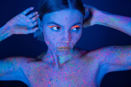 junge Frau in leuchtendem Neon-Make-up und farbenfroher Körperfarbe, die isoliert auf dunkelblauem Grund wegschaut