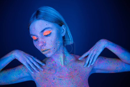 hübsche Frau in hellem Make-up und leuchtender Neon-Körperfarbe posiert isoliert auf dunkelblau