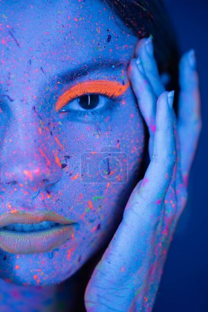 Photo for Close up portrait of cropped woman touching face with neon makeup and vibrant paint splashes isolated on dark blue - Royalty Free Image