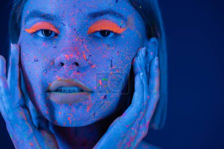 portrait of woman in vibrant neon makeup and body paint touching face and looking at camera isolated on dark blue