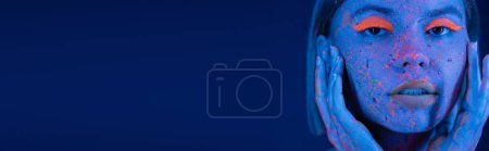 Téléchargez les photos : Portrait de jeune femme touchant le visage avec maquillage fluo et éclaboussures de peinture fluorescentes isolées sur bleu foncé, bannière - en image libre de droit