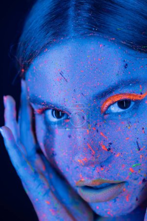 Photo for Close up portrait of woman with neon makeup and colorful paint on face looking at camera isolated on dark blue - Royalty Free Image