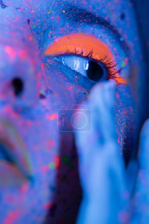 Nahaufnahme Porträt einer beschnittenen Frau mit hellem korallenfarbenen Lidschatten und bunten Farbflecken in blauem Neonlicht 