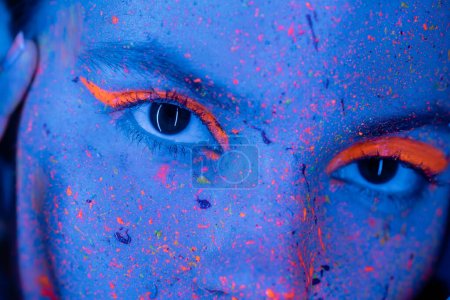 close up view of cropped woman with neon eye shadow and vibrant paint splatters looking at camera in blue light
