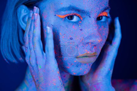 Photo for Portrait of woman in neon makeup and fluorescent paint holding hands near face on dark blue - Royalty Free Image