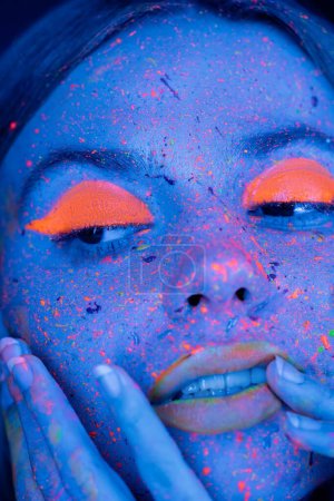 Foto de Primer plano retrato de la mujer con el maquillaje fluorescente tocando la cara en luz de neón azul - Imagen libre de derechos