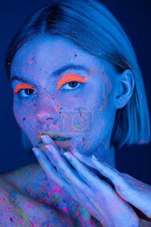 Téléchargez les photos : Portrait de femme au néon lumineux maquillage et peinture corporelle colorée touchant les lèvres isolées sur bleu foncé - en image libre de droit