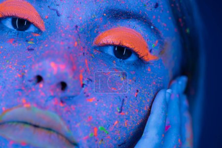 Nahaufnahme einer beschnittenen Frau, die Gesicht mit Neon-Make-up berührt und helle Farbspritzer in blauem Licht