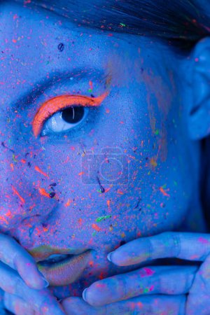 Foto de Vista parcial de la mujer cogida de la mano cerca de la cara con maquillaje de neón y salpicaduras de pintura brillante en luz azul - Imagen libre de derechos