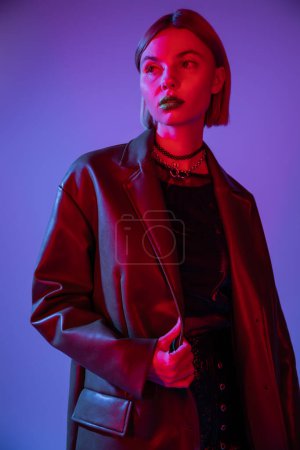 Téléchargez les photos : Femme à la mode en veste et colliers en cuir regardant loin dans la lumière rouge sur fond bleu - en image libre de droit