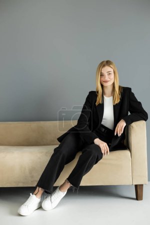 Smiling blonde woman in jeans and black jacket sitting on couch at home 
