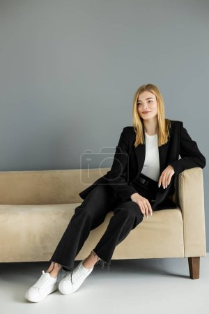 Fröhliche blonde Frau in Jeans und Jacke sitzt auf Couch neben grauer Wand 