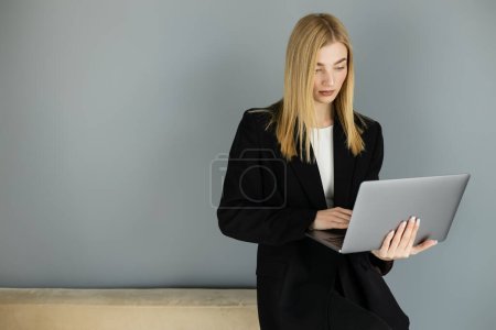 Téléchargez les photos : Autocar blonde utilisant un ordinateur portable près du canapé et du mur gris à la maison - en image libre de droit