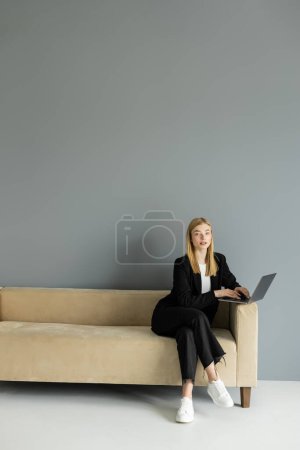 Coach blonde utilisant un ordinateur portable et regardant la caméra tout en étant assis sur le canapé à la maison 
