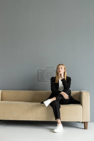 Femme pensive en veste assise sur un canapé beige à la maison 