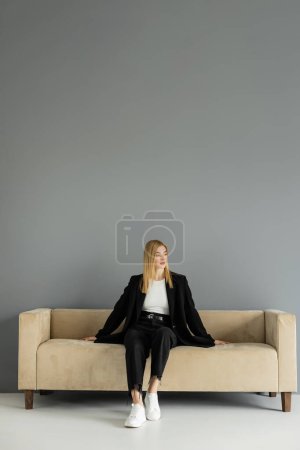Junge blonde Frau in schwarzer Jacke sitzt zu Hause auf beiger Couch 