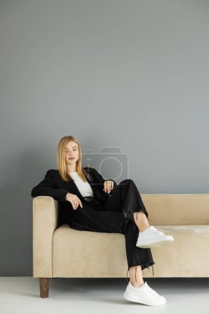 Pretty blonde woman in jacket looking at camera while resting on couch at home 