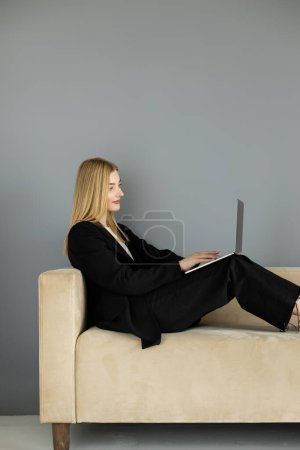 Smiling coach in jacket using laptop on sofa near grey wall 