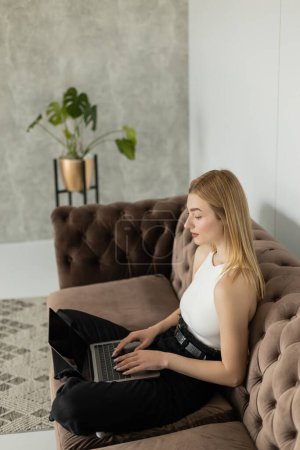 Young woman using laptop with blank screen while sitting on brown couch at home 