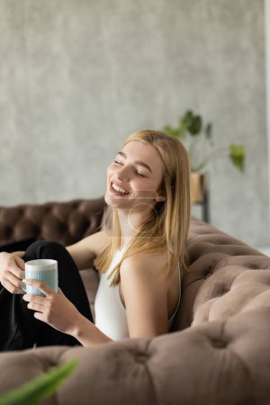 Young blonde woman laughing and holding cup while sitting on couch at home 