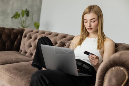 Blonde Frau im Top mit Kreditkarte und Laptop auf bequemer Couch 