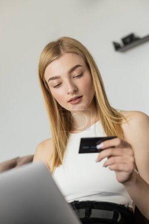 Téléchargez les photos : Jeune femme regardant la carte de crédit floue près d'un ordinateur portable à la maison - en image libre de droit