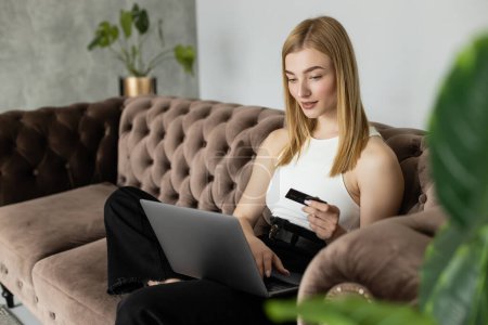 Positive blonde woman holding credit card and using laptop on modern couch near plants at home 