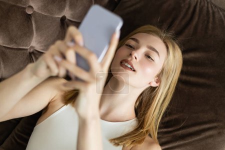 Vista superior de la mujer rubia sonriente usando un teléfono inteligente borroso en el sofá 