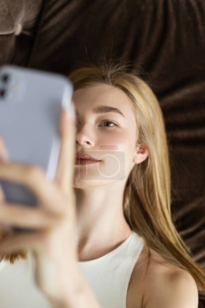 Top view of young woman in top using blurred cellphone on couch 