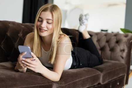 Photo for Pleased blonde woman using mobile phone while relaxing on couch at home - Royalty Free Image