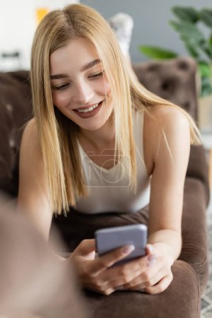 Joyful woman in top using blurred smartphone while relaxing on couch 