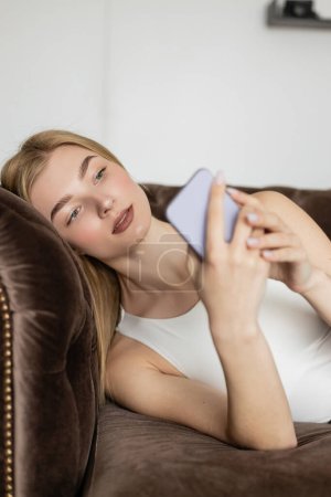 Jolie jeune femme regardant flou téléphone portable sur canapé brun à la maison 