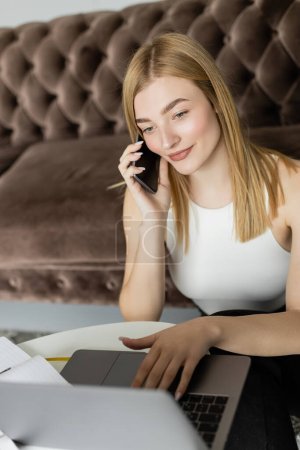 Téléchargez les photos : Jeune coach blonde utilisant un ordinateur portable et parlant sur smartphone près d'un ordinateur portable à la maison - en image libre de droit