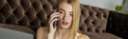 Photo for Blonde coach talking on smartphone and looking at blurred laptop near sofa at home, banner - Royalty Free Image