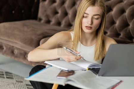 Mujer joven mirando portátil cerca de dispositivos durante la educación en línea en la sala de estar 