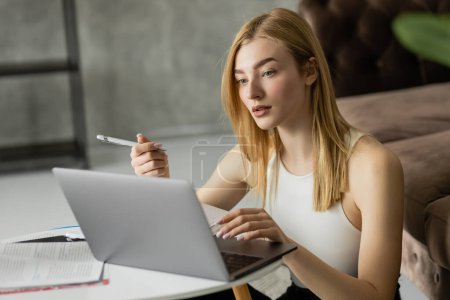 Mujer joven sosteniendo la pluma y el uso de portátil cerca de portátil durante la educación en línea 