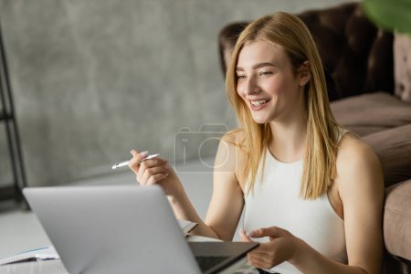 Blonde woman smiling while holding pen near laptop and notebooks during online education 