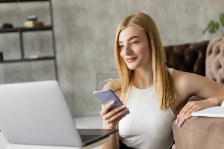 Positive coach holding smartphone near blurred laptop while working at home 
