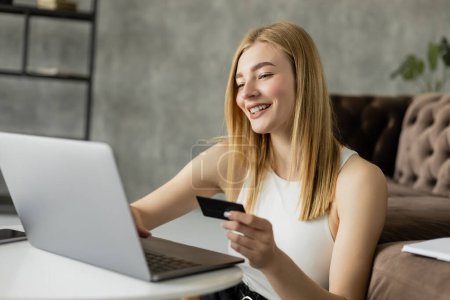 Mujer rubia positiva usando portátil y tarjeta de crédito en la sala de estar 