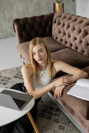 Portrait of blonde coach looking at camera near laptop and notebook at home 