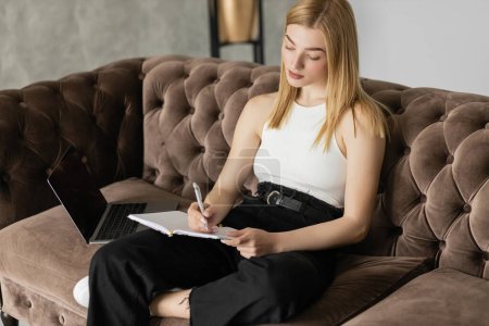 Mujer rubia joven escribiendo en el cuaderno durante el aprendizaje electrónico en el portátil en el sofá 