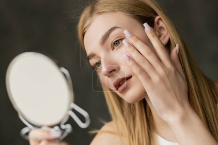 Young blonde woman touching face and holding blurred mirror at home 