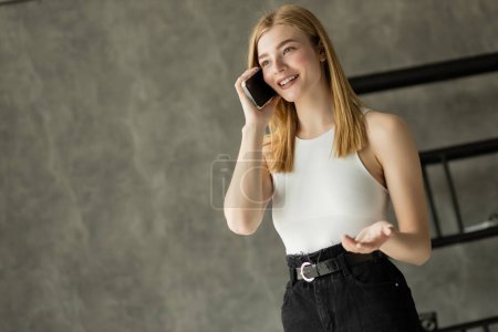 Foto de Mujer rubia positiva en la parte superior hablando en el teléfono inteligente en casa - Imagen libre de derechos