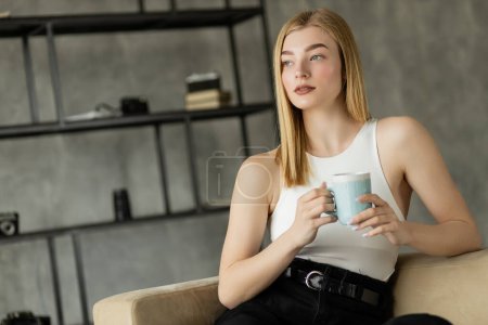 Young blonde woman holding coffee cup while sitting on couch in living room 