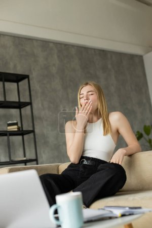 Blonde freelancer yawning near blurred gadgets and coffee in living room 