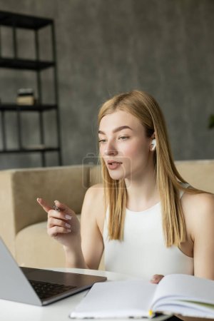 Blonde Trainerin im Kopfhörer zeigt beim Videoanruf am heimischen Laptop mit dem Finger 