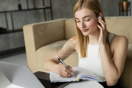 Blonde woman listening music in earphone and writing on notebook near laptop during online education at home 