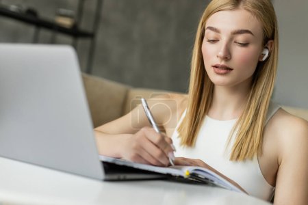 Blonde Frau mit Kopfhörer schreibt beim E-Learning in der Nähe von Laptop zu Hause auf Notizbuch 